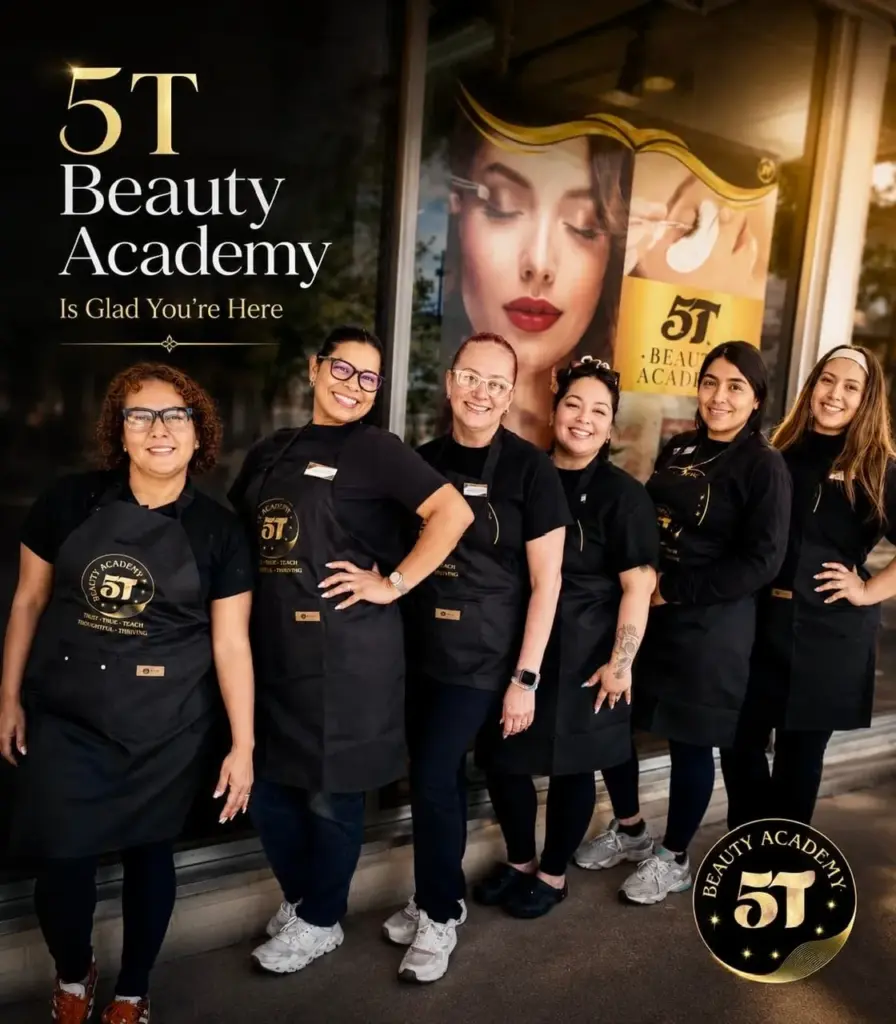 A group of six smiling female beauty instructors wearing black aprons with the 5T Beauty Academy logo, standing in front of the academy's storefront window.