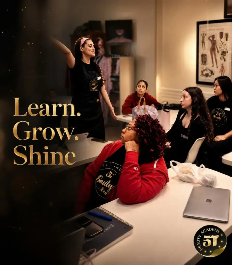 An instructor with a pink headband gesturing during a lecture to a classroom of attentive beauty school students seated at desks with laptops.